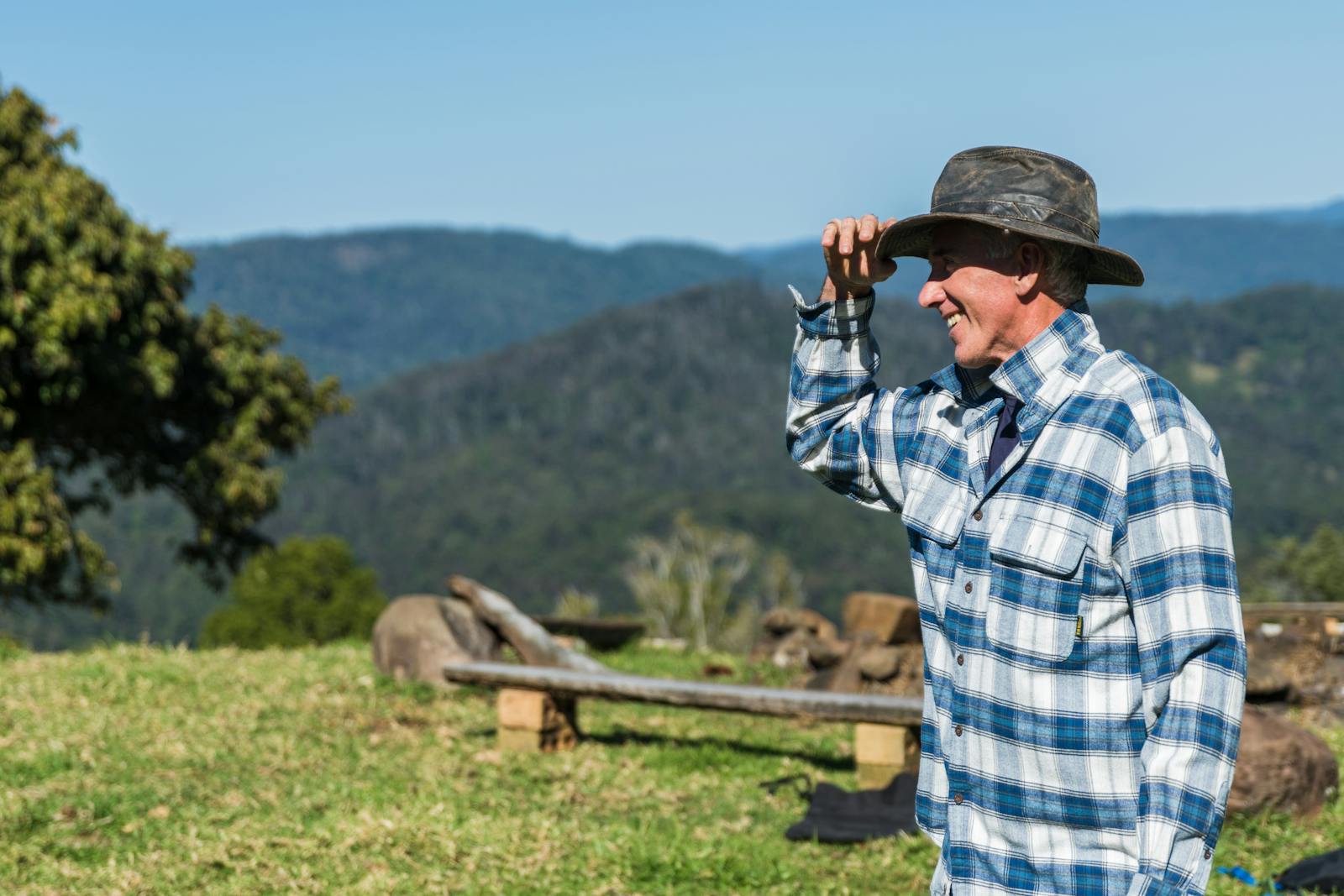 A smiling farmer in plaid flannel enjoys a sunny day on a lush green hillside.